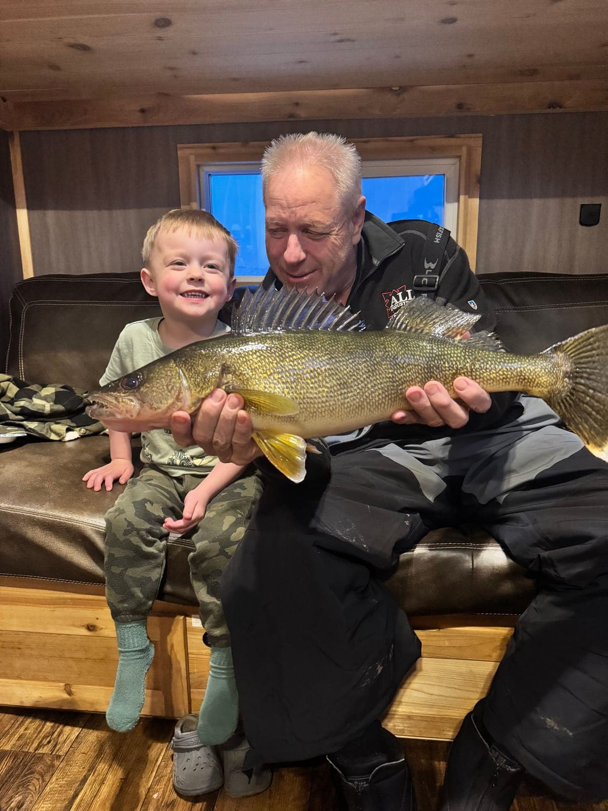A man and a young boy sitting on a couch inside a fish house, holding up a big walleye
